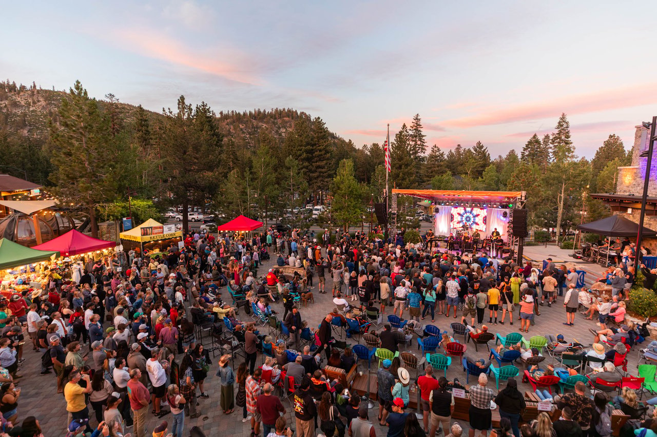 Crowd watches an outdoor concert at dusk, with vendor tents and pine-covered hills behind the stage.