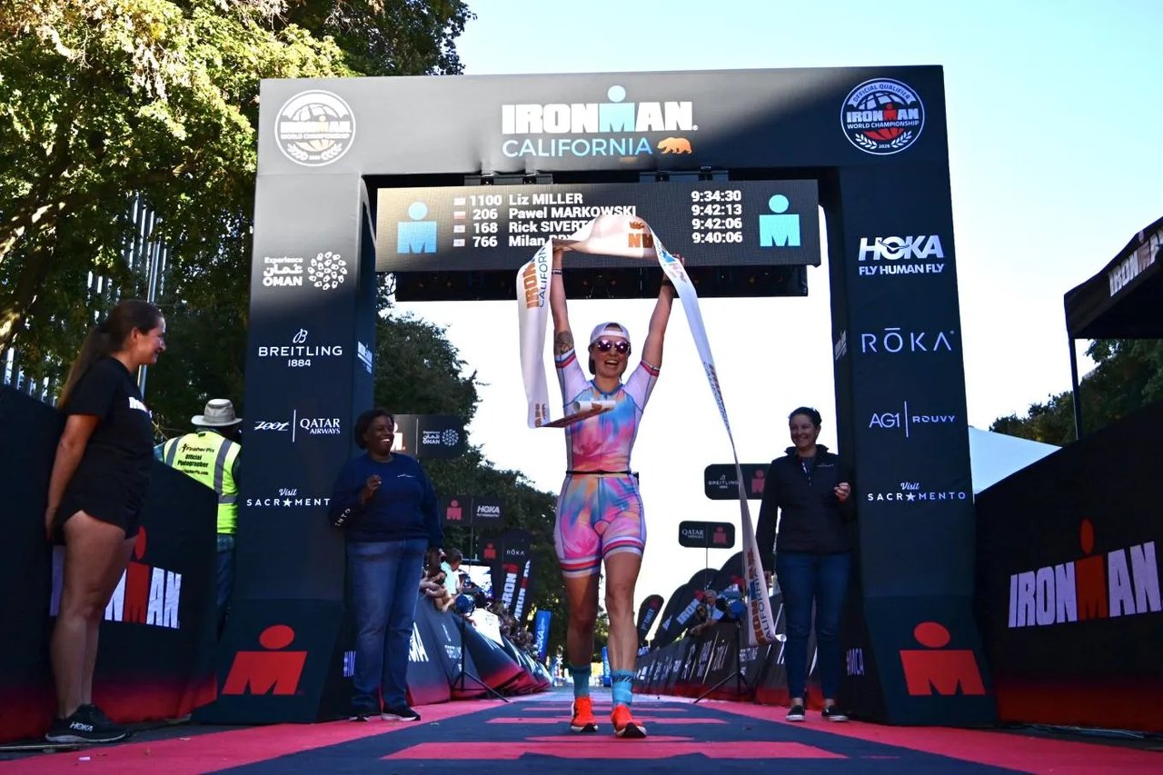 Triathlete raises a finisher's tape beneath the IRONMAN California arch in Sacramento.