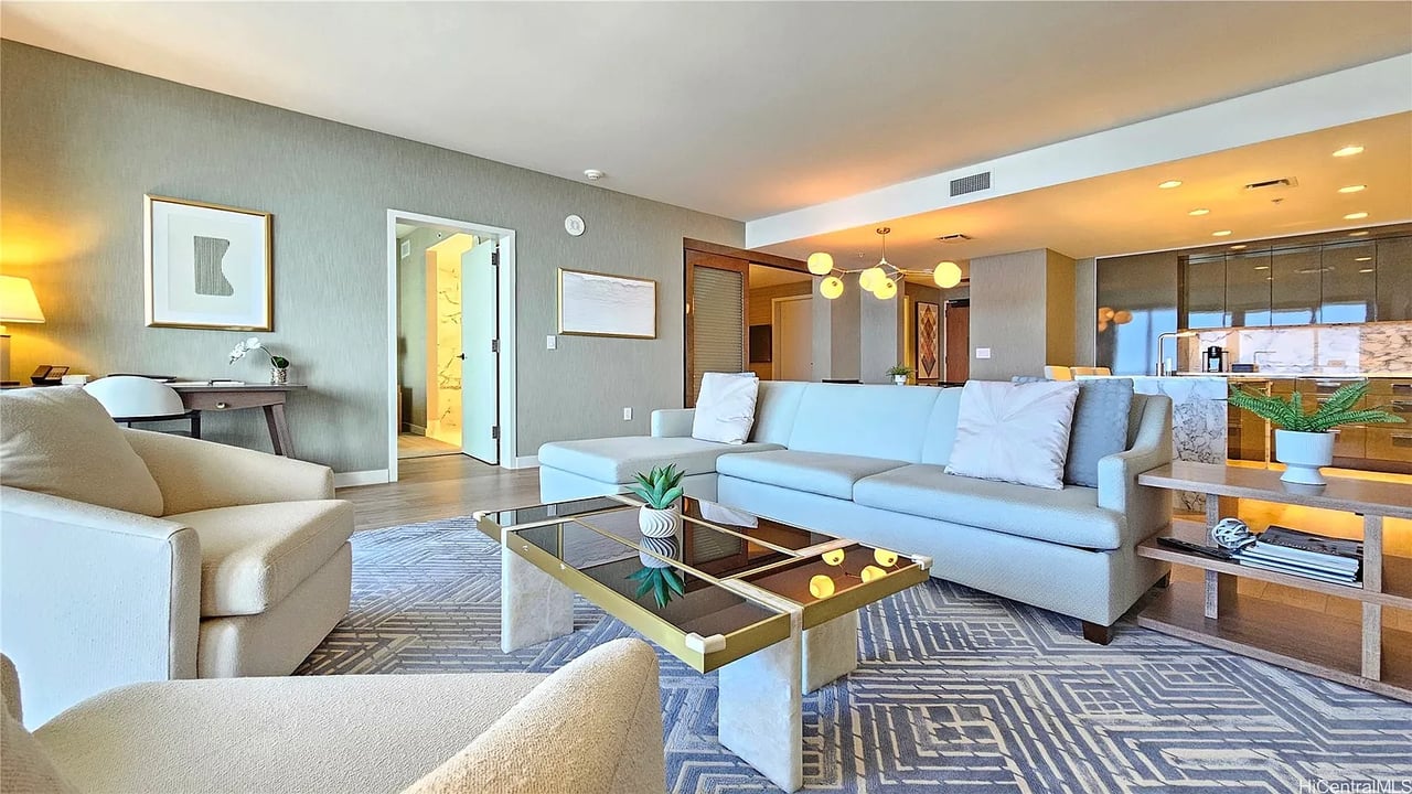 Furnished living room with pale sofas, a glass coffee table, and an open kitchen with a marble island.