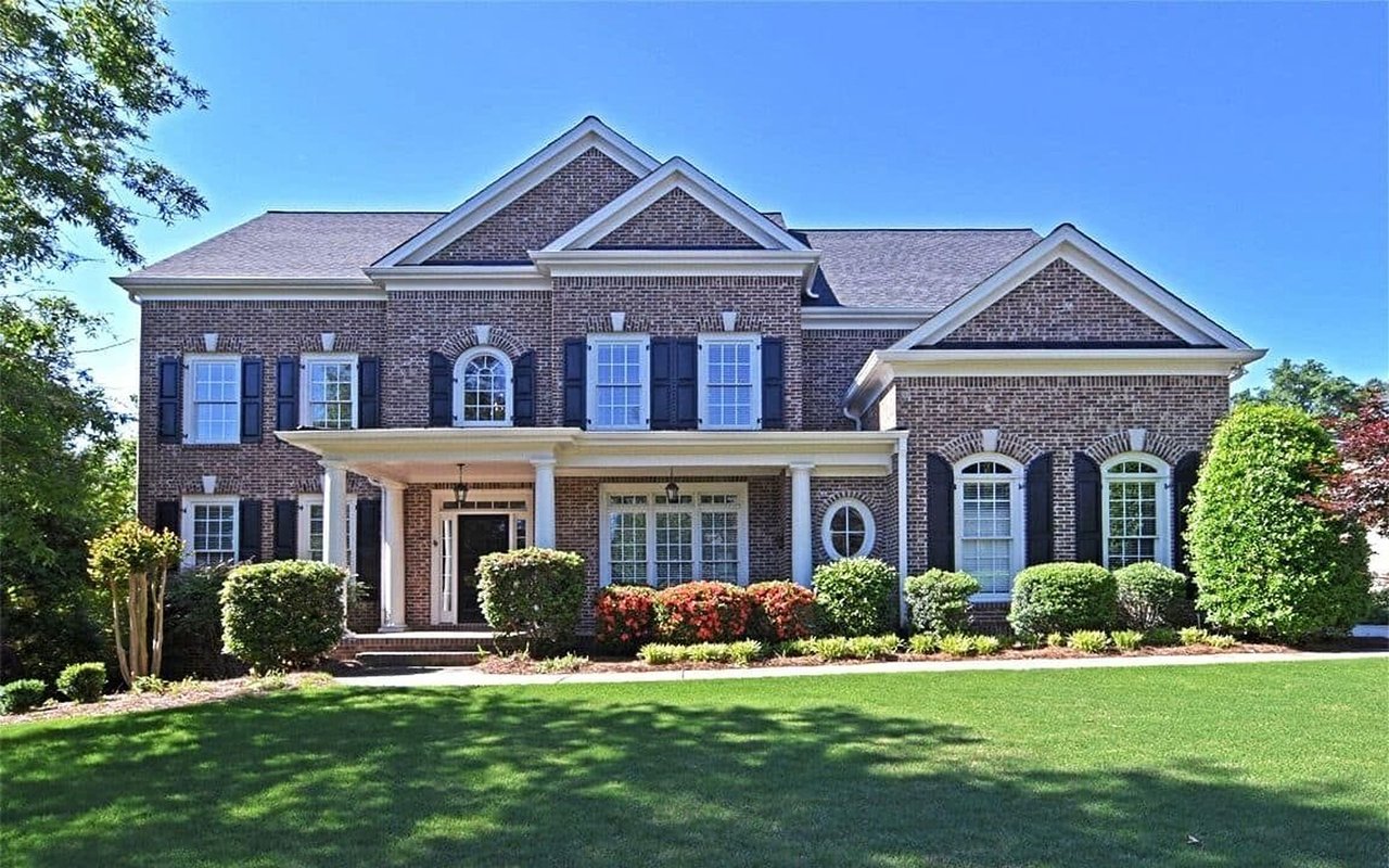 Two-story brick house with a covered porch, dark shutters, and landscaped lawn.