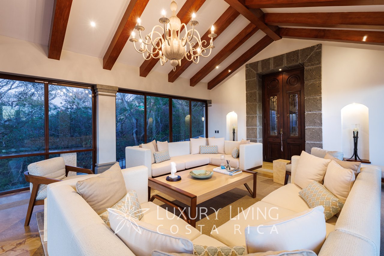 Furnished living room with exposed timber beams, a chandelier, and large windows overlooking trees.
