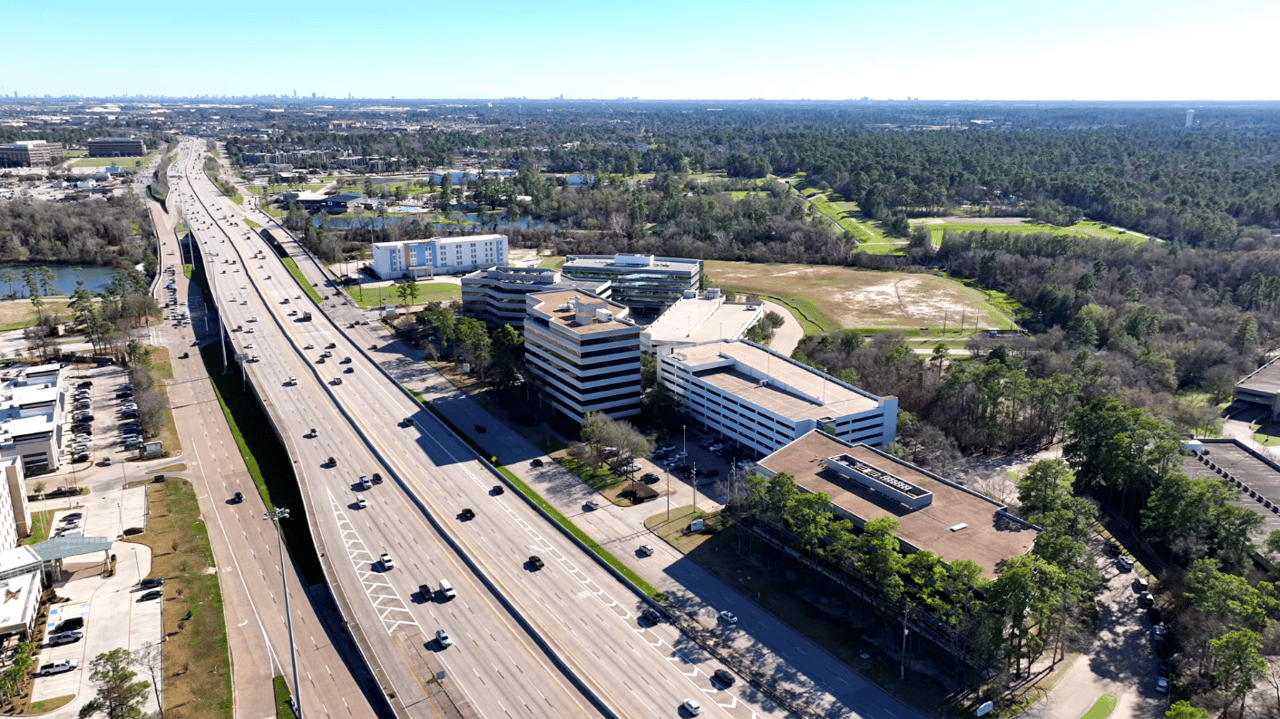 Commercial office park beside a multi-lane highway, with modern buildings and wooded surroundings.