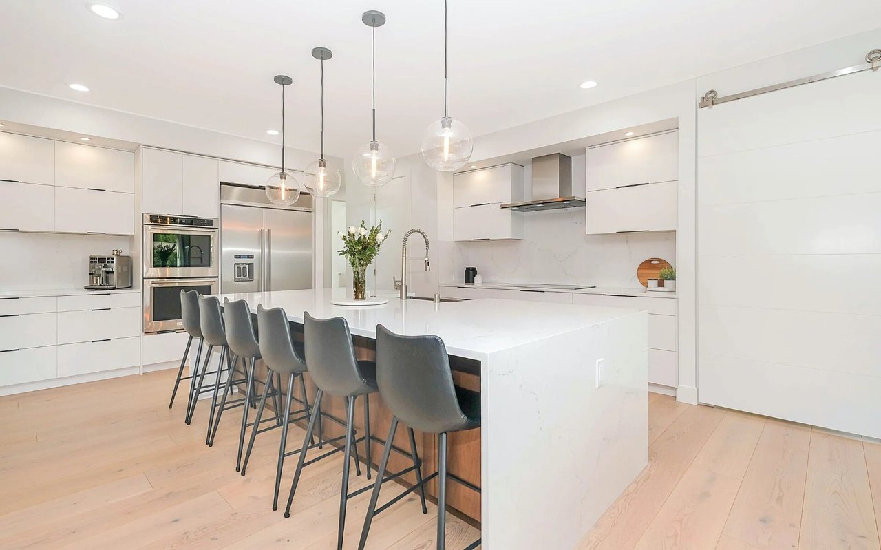 Modern white kitchen with a large island, gray bar stools, and stainless steel appliances.