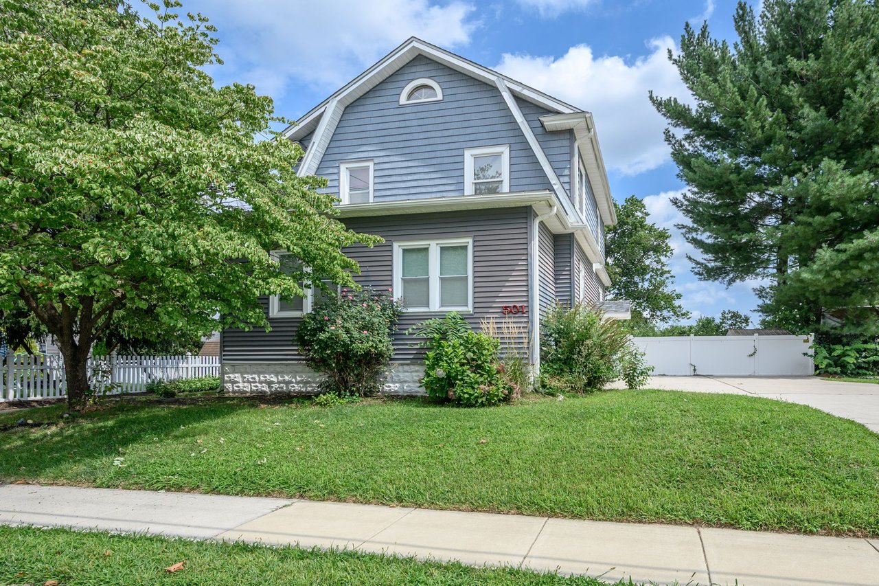 Blue-gray house with a gambrel roof, white picket fence, and mature trees.