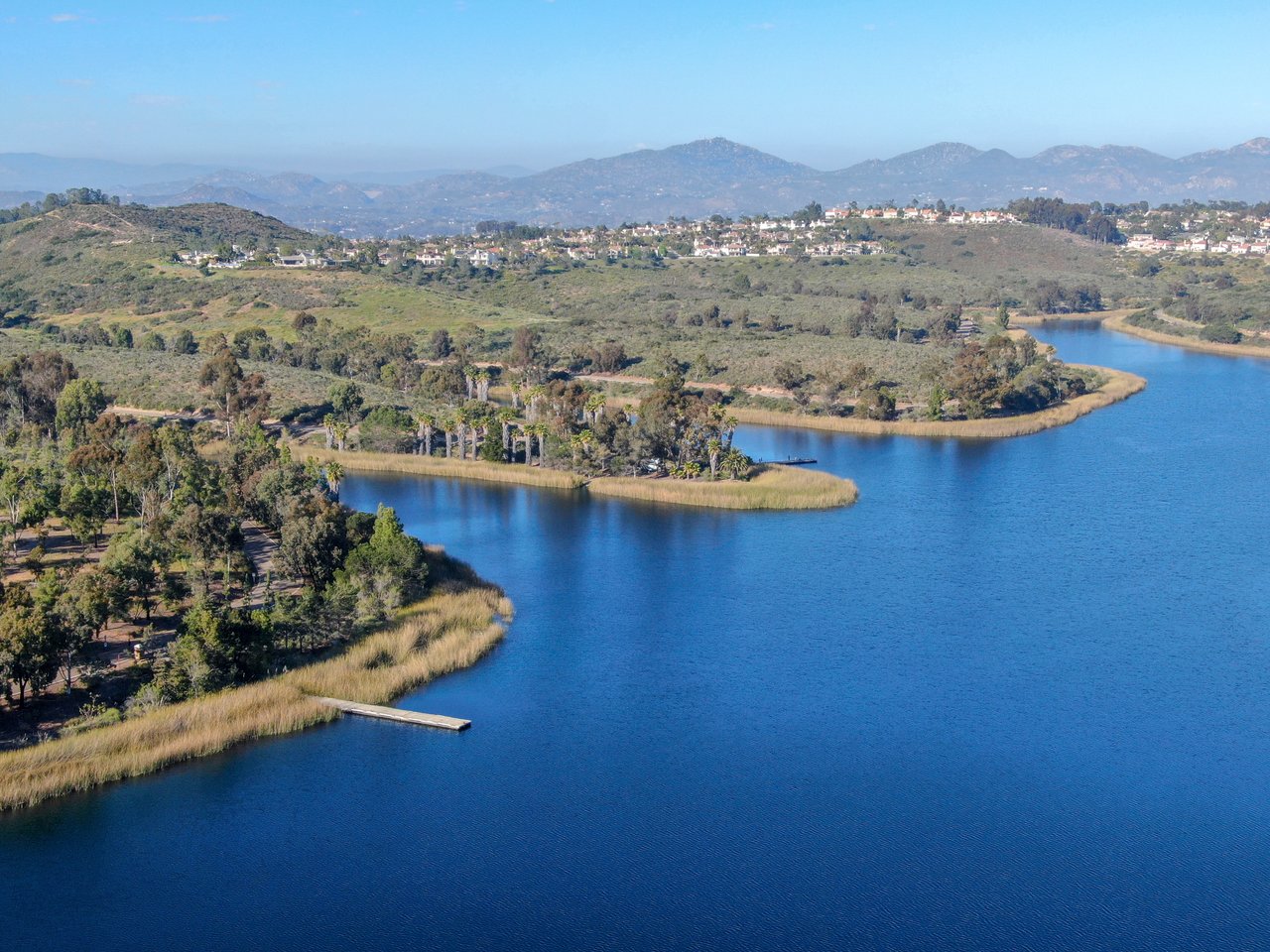 Aerial view of a blue lake, marshy shoreline, wooded parkland, and homes among distant hills in Scripps Ranch.