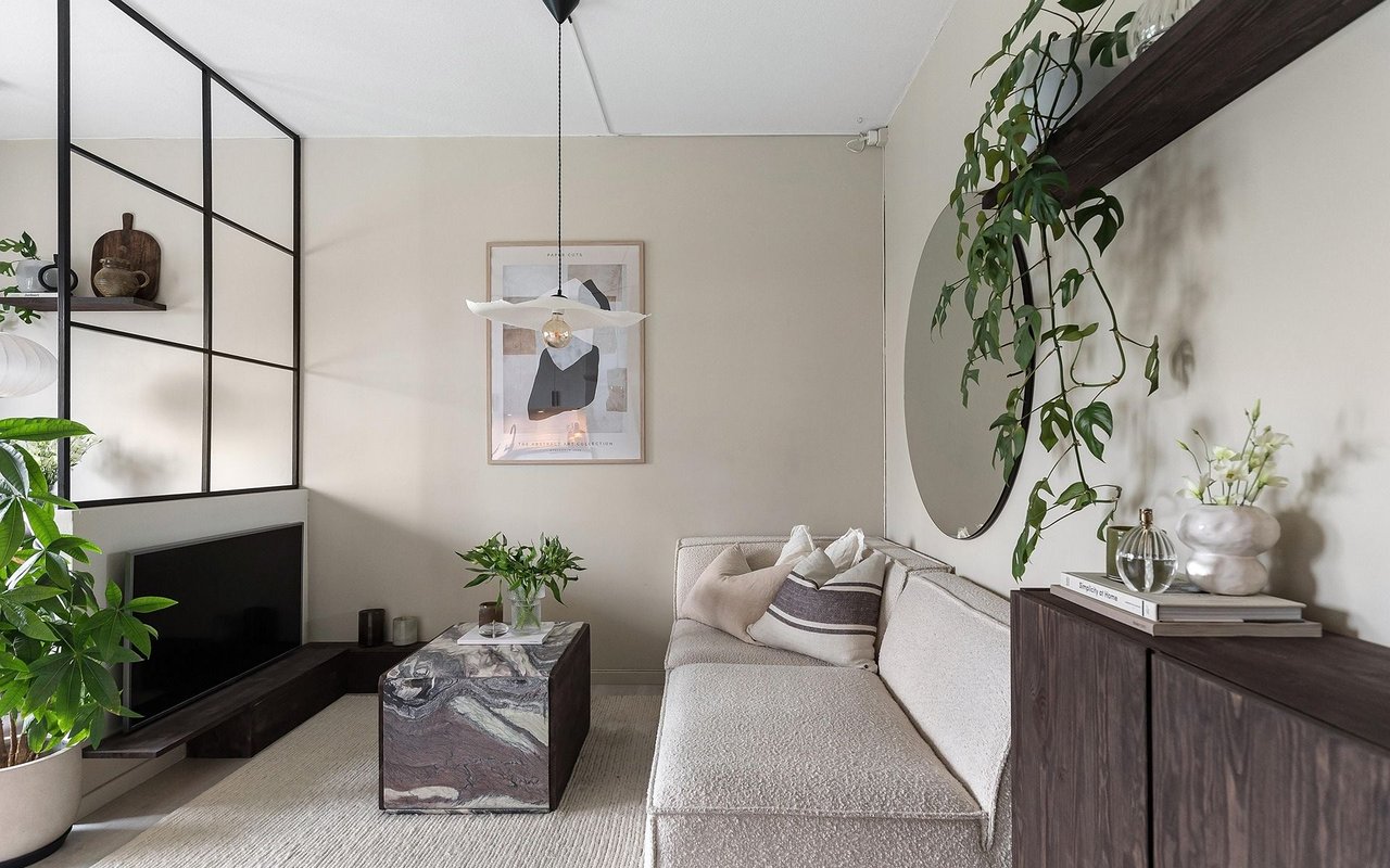 Living room with a cream sectional, marble coffee table, round mirror, and leafy plants.
