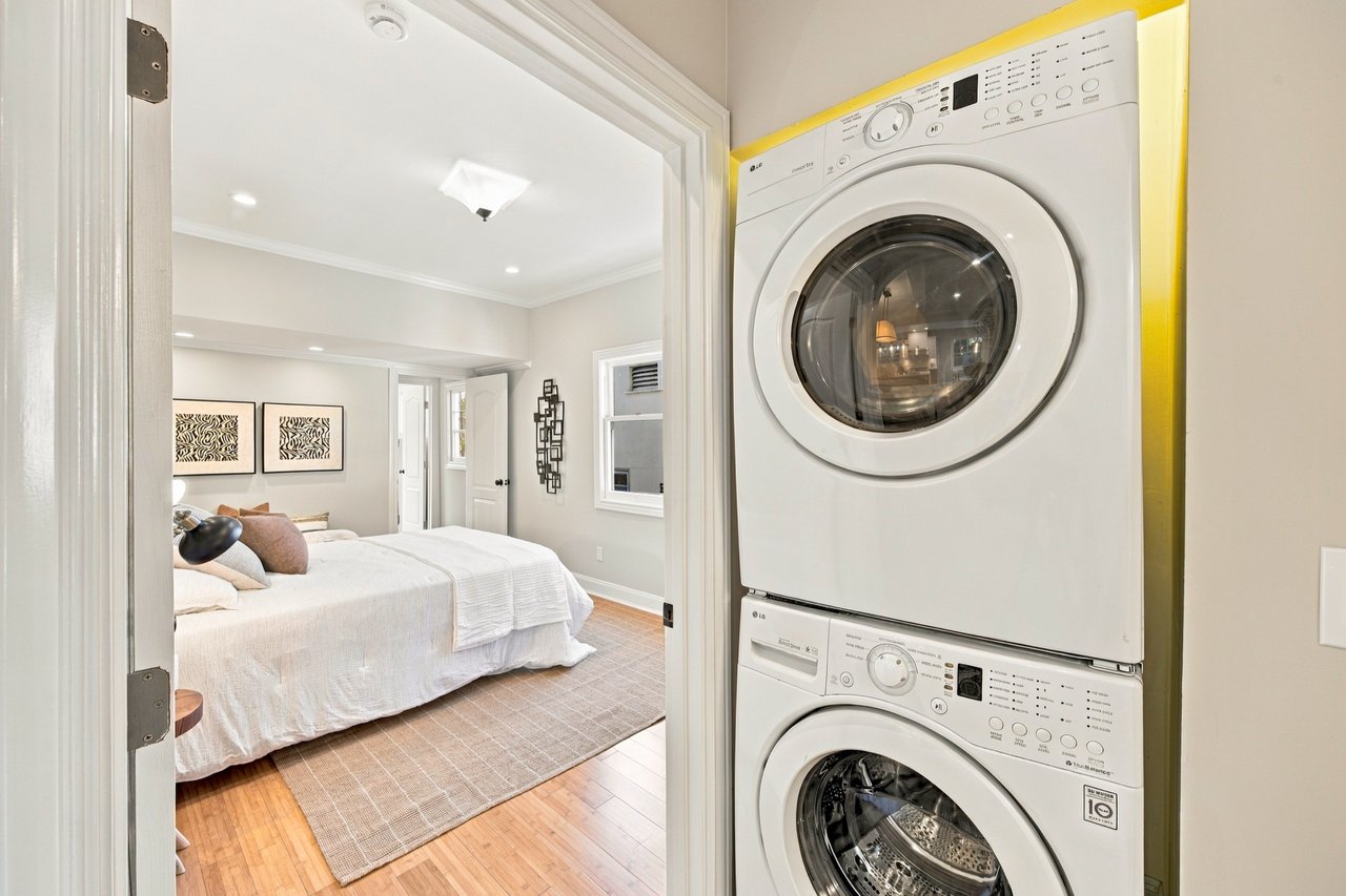 Stacked washer and dryer beside a furnished bedroom with hardwood floors.