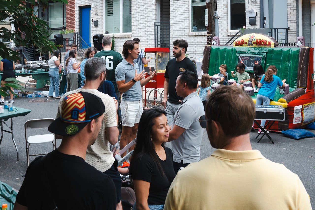 Neighbors mingle at a street block party as children play a colorful Zap-A-Mole game.