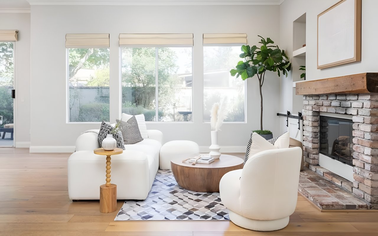 A bright living room with a white sectional sofa, a round wooden coffee table, and a brick fireplace with a wood mantle.