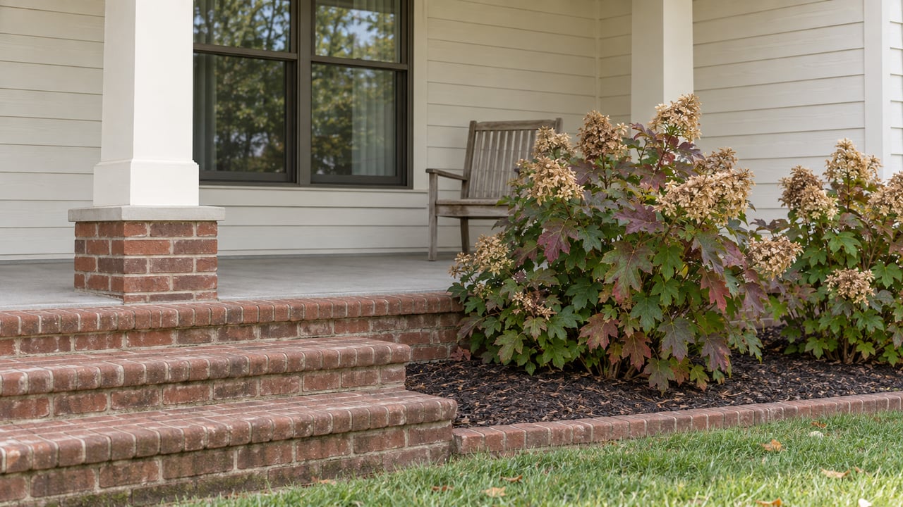 Brick porch steps beside oakleaf hydrangeas, with cream wood siding, a bronze-framed window and a weathered wooden chair.