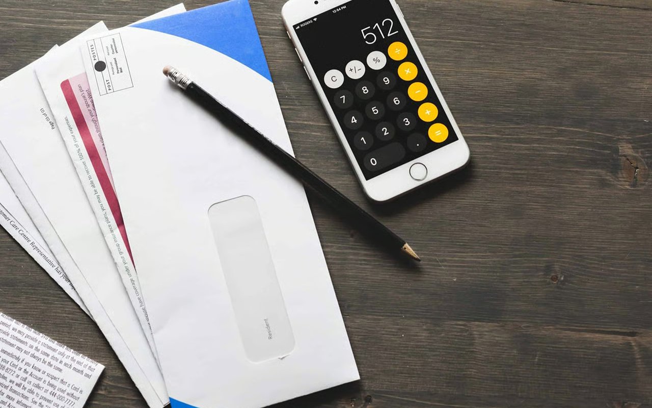 Smartphone calculator showing 512 beside a pencil, envelopes, and paperwork on a dark wood surface.