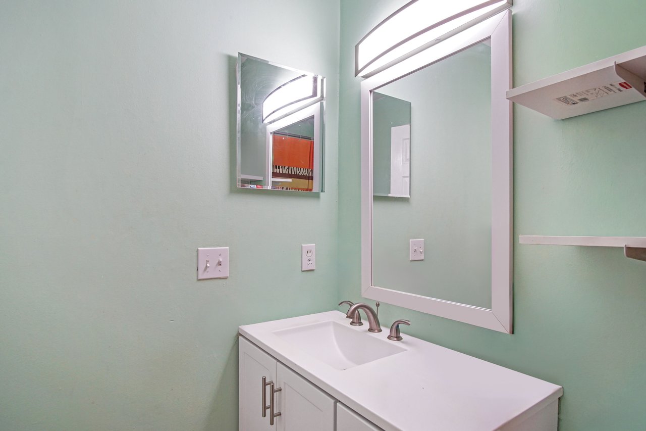 Bathroom at 2506 Nugget Lane in Tallahassee, Florida, featuring a white vanity with storage, modern brushed-nickel faucet, and large framed mirror with overhead light. Walls are painted a soft mint green, and shelving is mounted for additional storage.