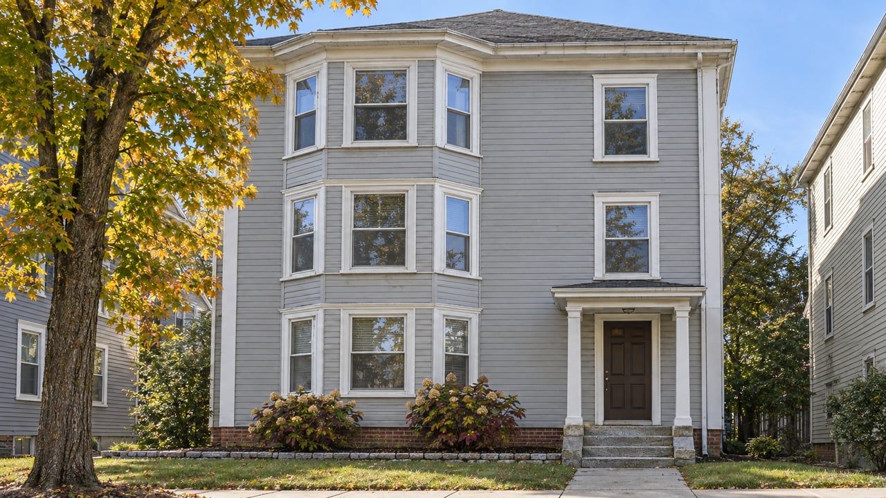 Three-story light gray clapboard home with stacked bay windows, a covered entrance, granite steps and small autumn planting beds.