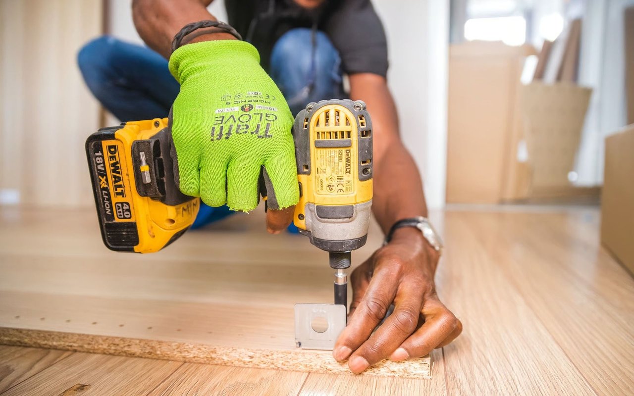 Worker uses a cordless drill to fasten a metal bracket to a board on a wood floor.