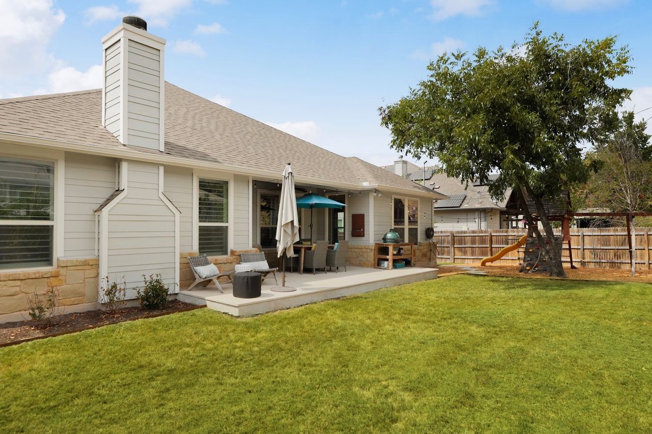 House rear patio with outdoor seating, a teal umbrella, green lawn, and a fenced yard with a playset.