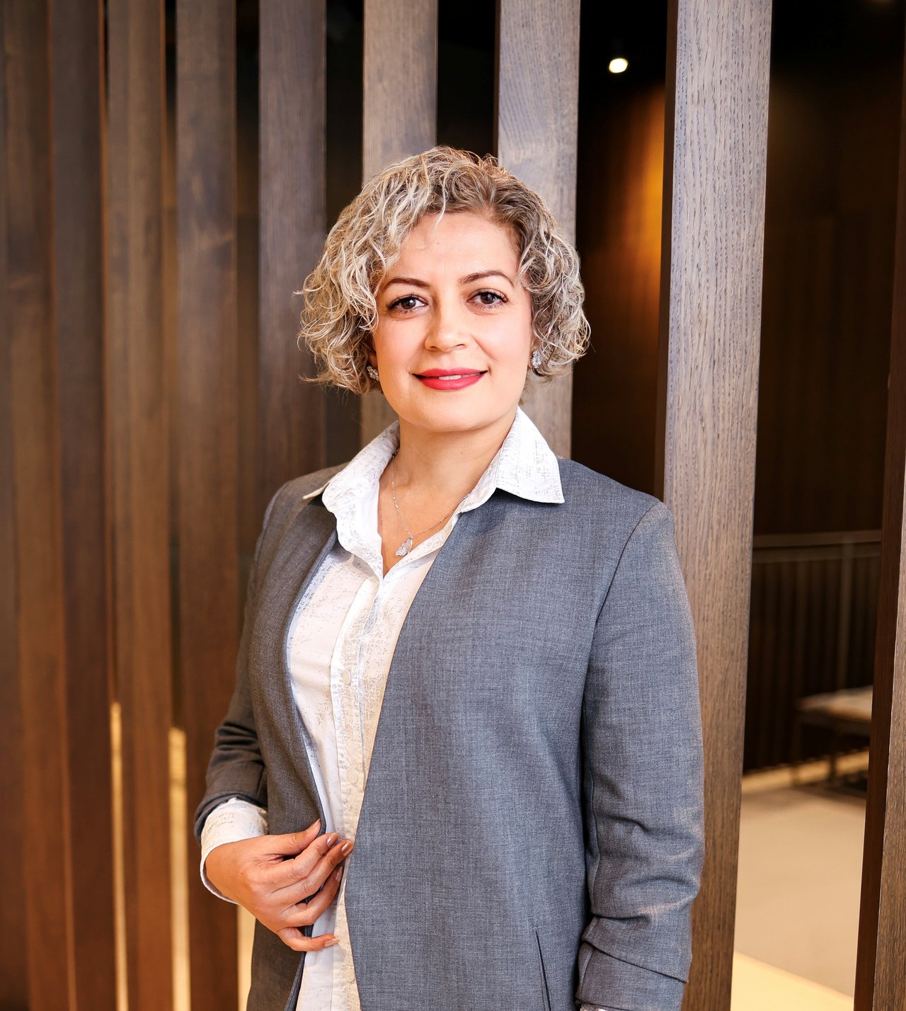 Ensieh Davatgar in a gray suit jacket and white blouse, standing before wooden panels.
