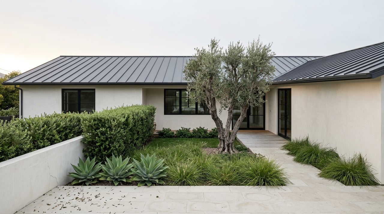 Low-roofed plaster home beyond a courtyard with an olive tree, agaves, and limestone terrace.
