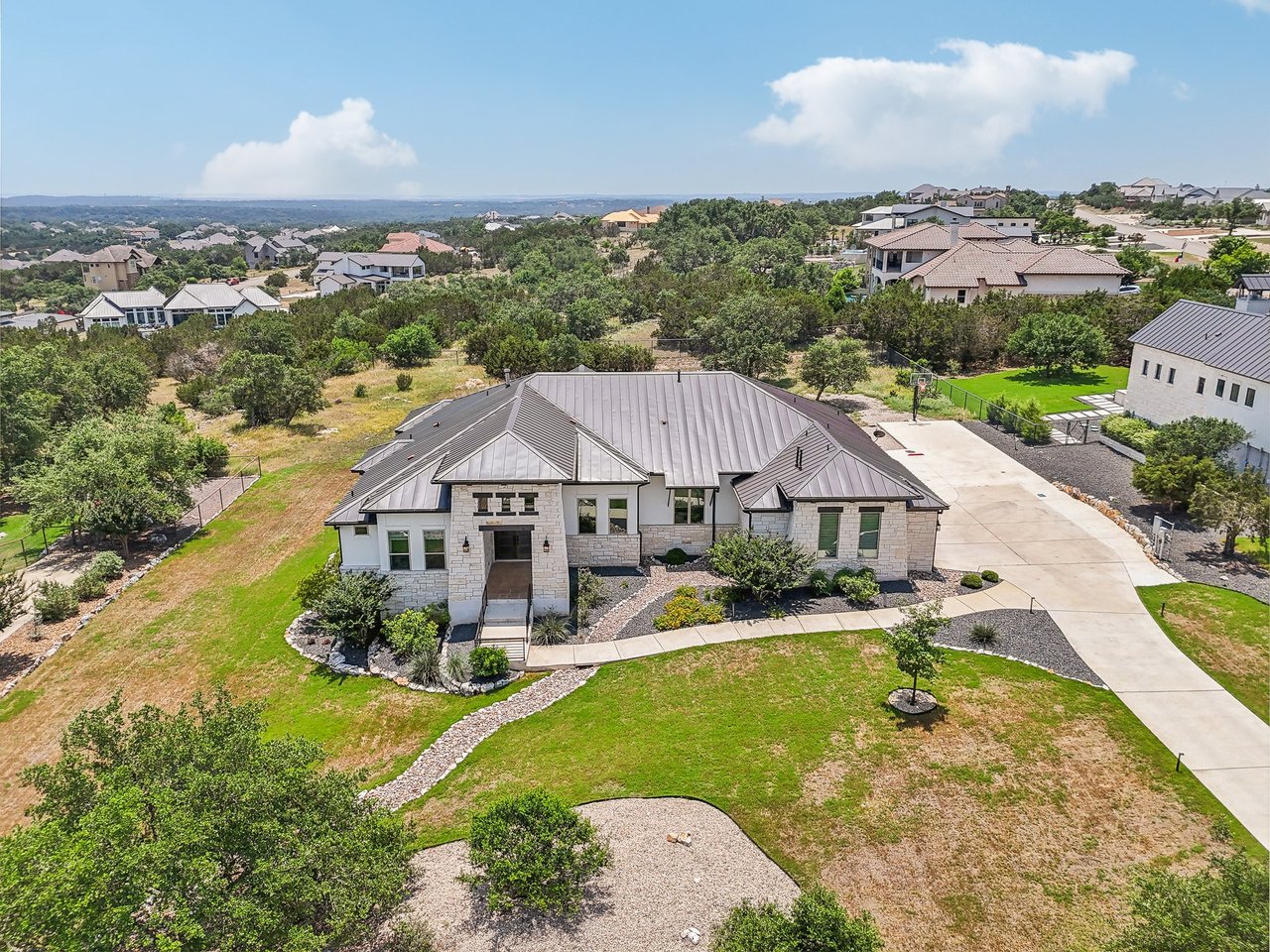 Aerial view of a luxury Texas hill country home. It has a metal roof with hardscape flower beds on a large 1 acre lot. There is a backdrop view of other homes with rolling hills and white puffy clouds in the sky.