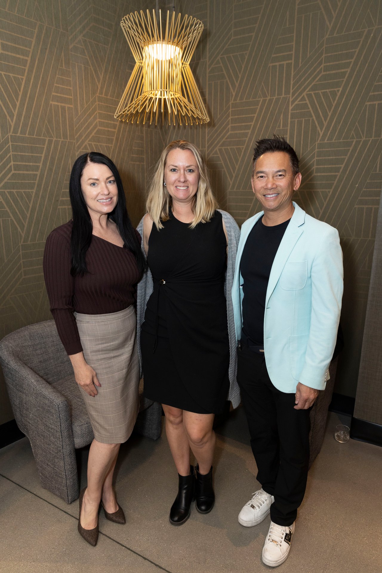Three adults pose together beside a gray chair beneath a gold wall light.