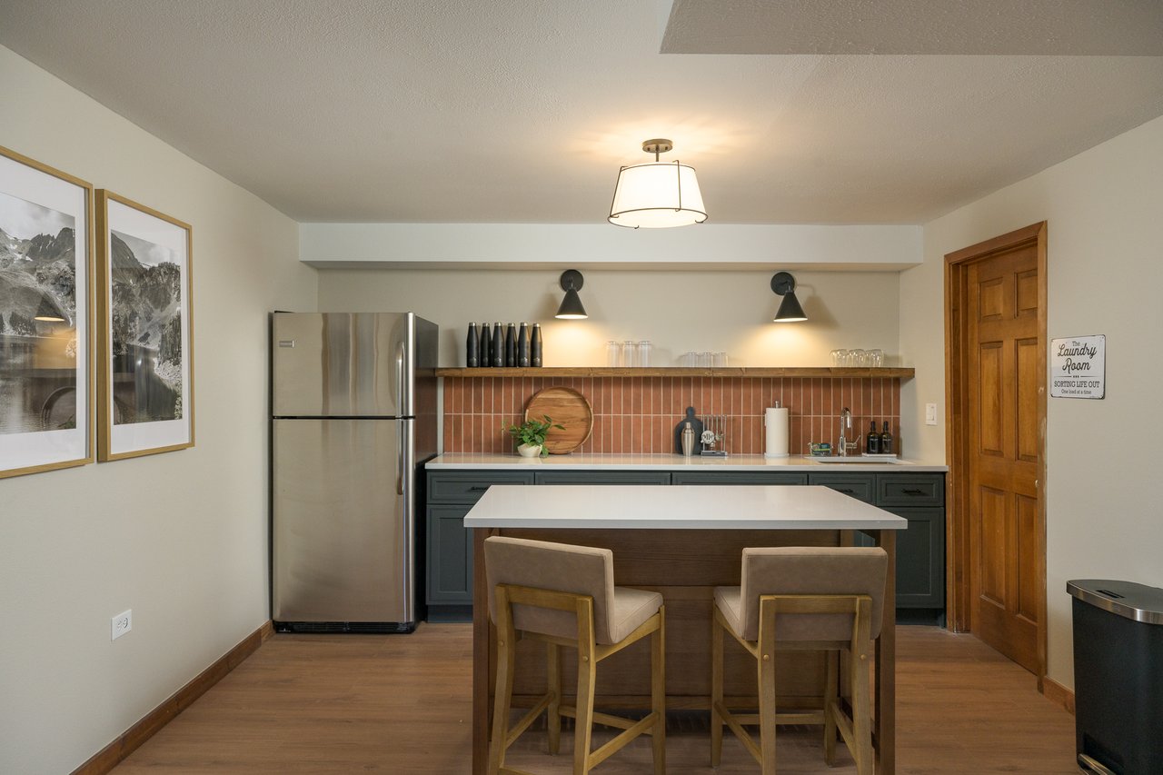 Kitchen with an island, two upholstered stools, dark green cabinets, and a terracotta tile backsplash.