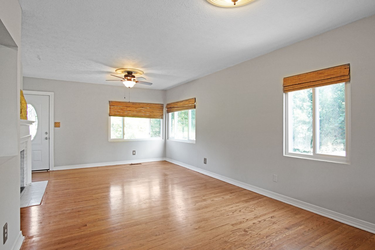 Bright, empty living room with light wood flooring, white walls, and a ceiling fan. Three windows with bamboo shades let in natural light, creating an open, airy feel.