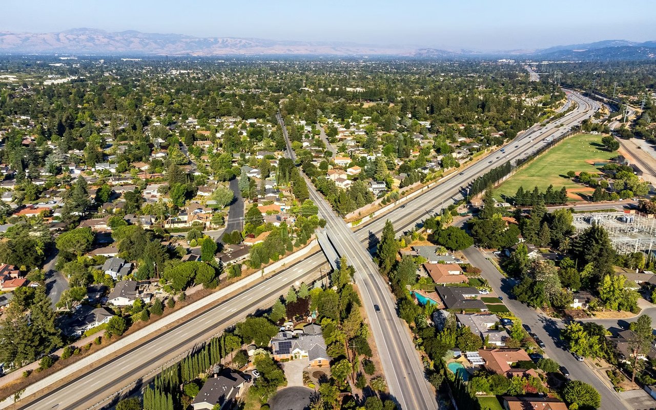 Aerial view of a leafy neighborhood with a highway interchange, sports fields, and distant hills.