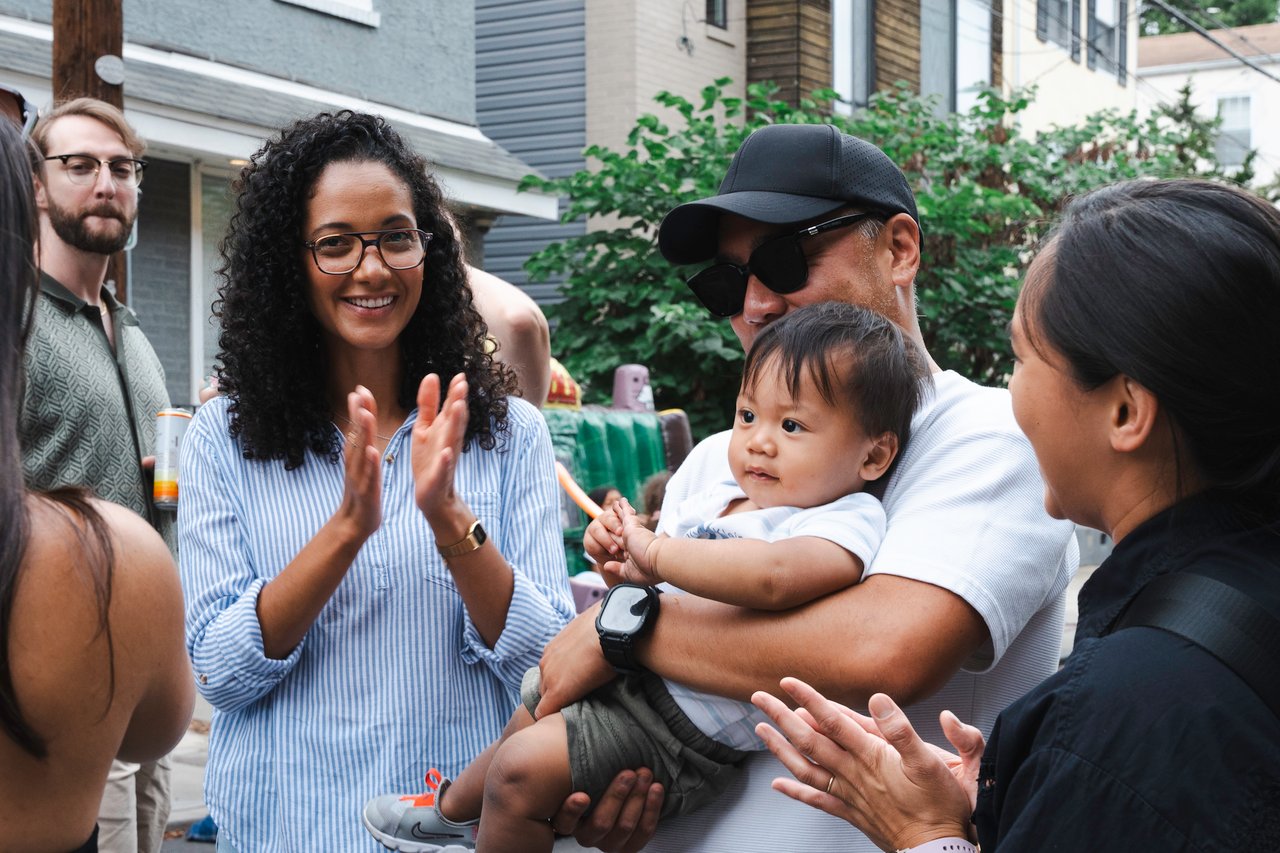 Neighbors gather at a block party as a woman claps and a man holds a baby.