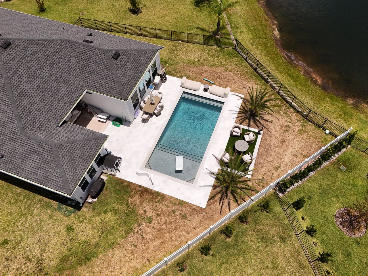 Aerial view of a home’s backyard with a rectangular pool, stone patio, and waterfront lawn.