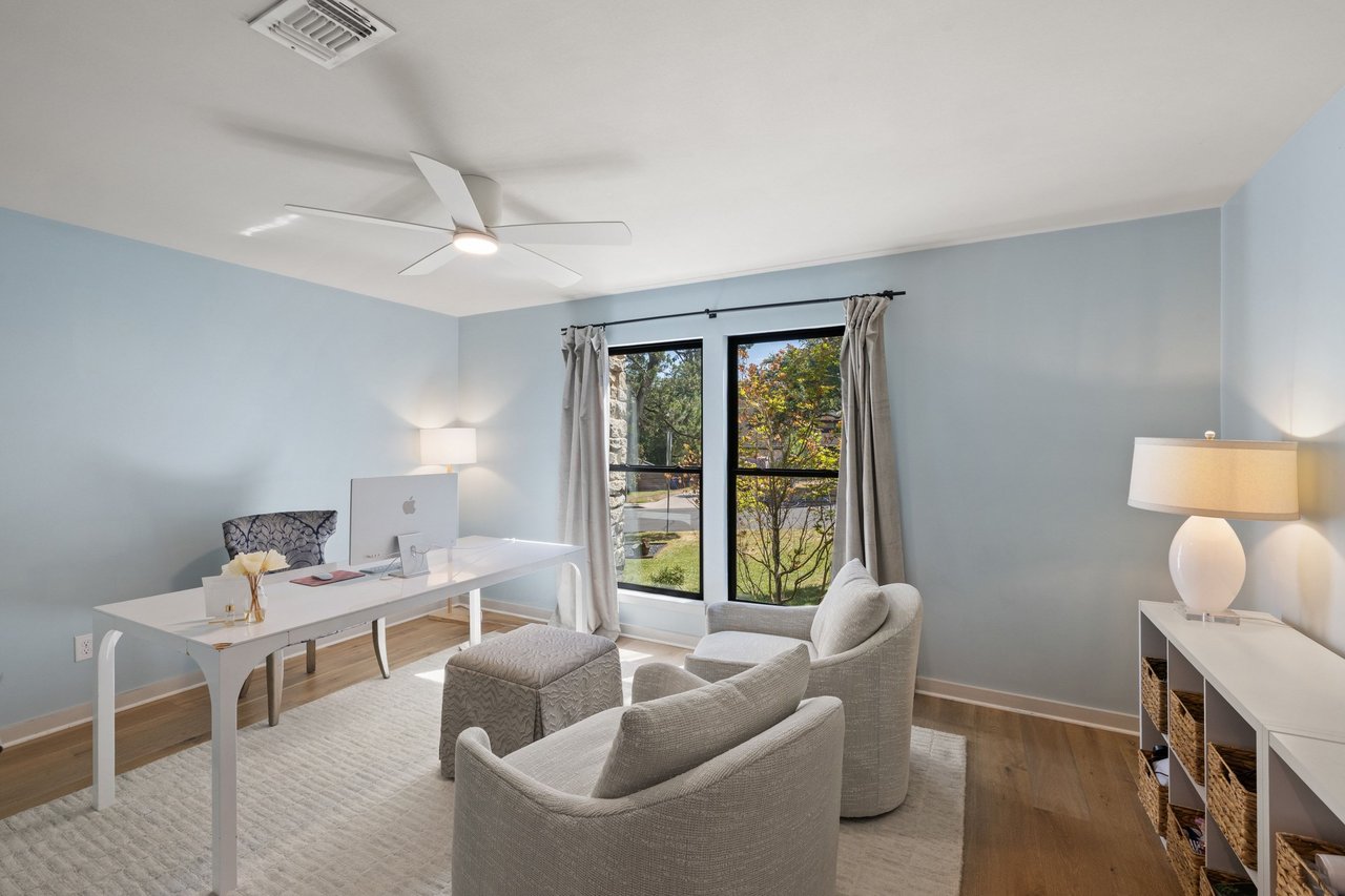 Staged home office with a white desk, upholstered chairs, and a large window overlooking the yard.