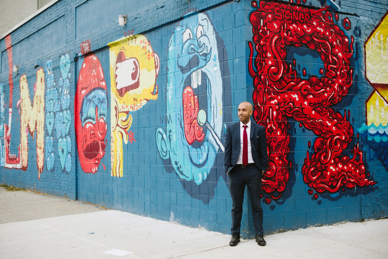 Man in a dark suit standing beside a colorful graffiti-covered wall.