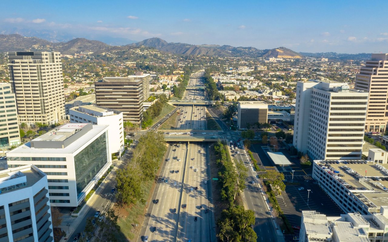 Aerial city view with a freeway between office buildings and hills in the distance.