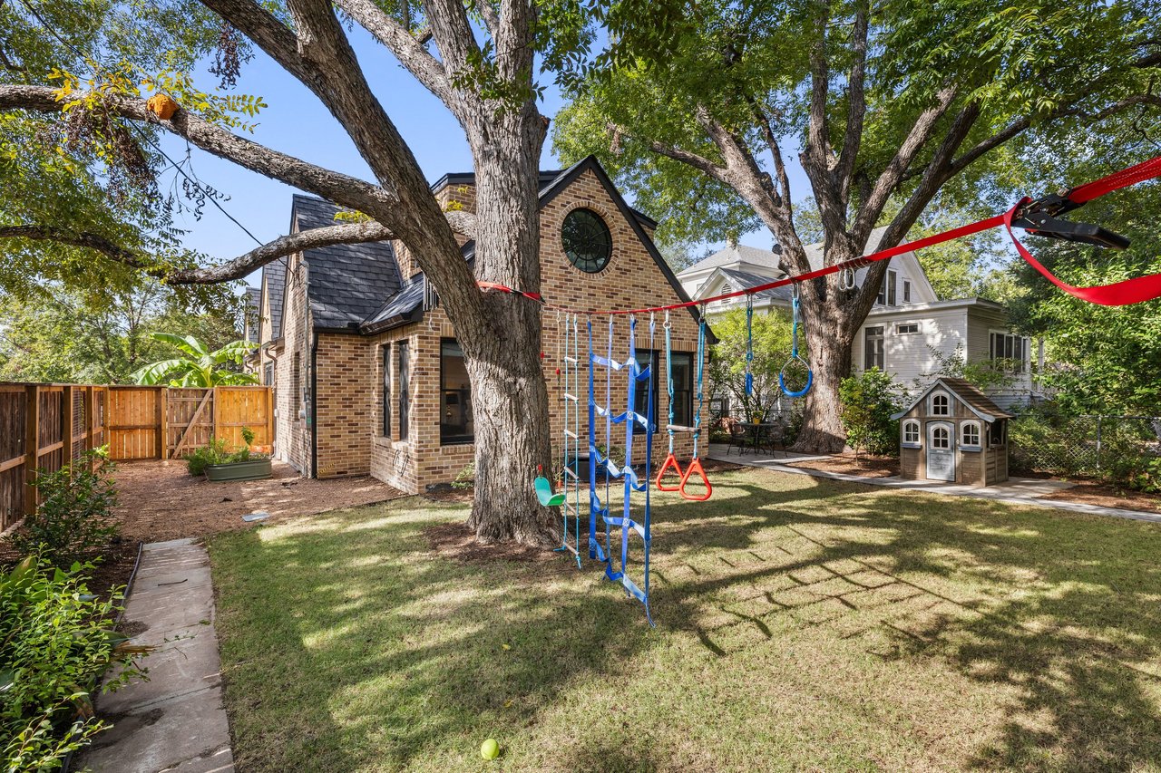 Backyard with a brick house, large shade trees, a swing set, and a small playhouse.