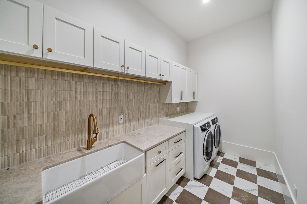 Laundry room with a farmhouse sink, brass faucet, tiled backsplash, and front-loading washer and dryer.