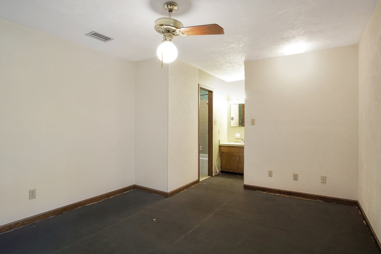 Empty room with beige walls and dark carpet. Ceiling fan with a light hangs overhead. An open doorway reveals a small bathroom with a sink and mirror.