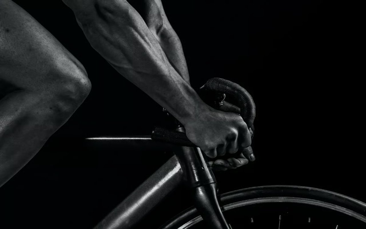 A black-and-white, close-up shot of a cyclist's hands gripping handlebars, with muscular arms and legs visible in profile.