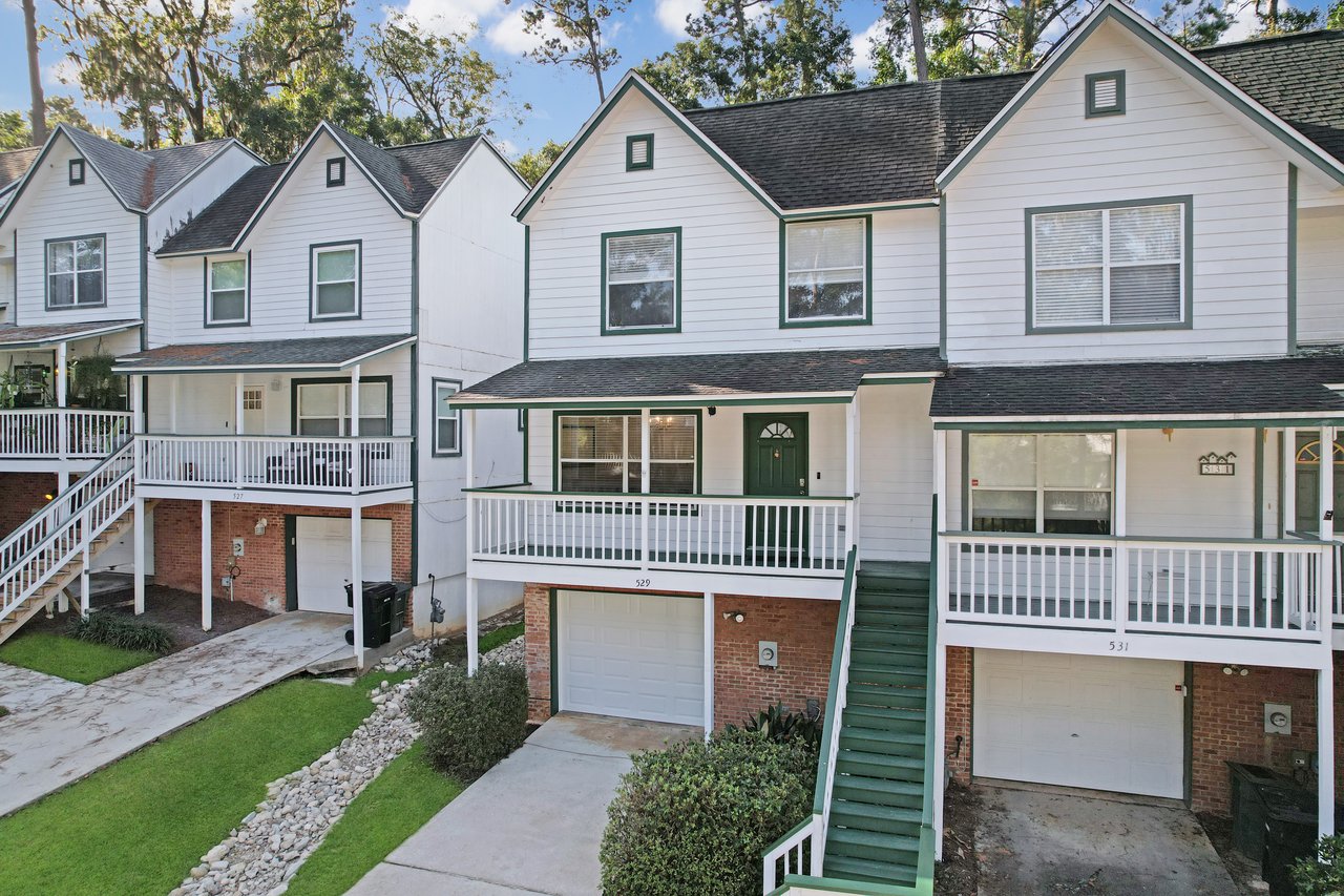 Front exterior of the townhome at 529 Collinsford Road, Tallahassee, Florida, showing a two-story design with white siding, green trim, and a one-car garage. The home features a covered front porch with white railings, a green staircase leading to the main entrance, and brick accents on the lower level.