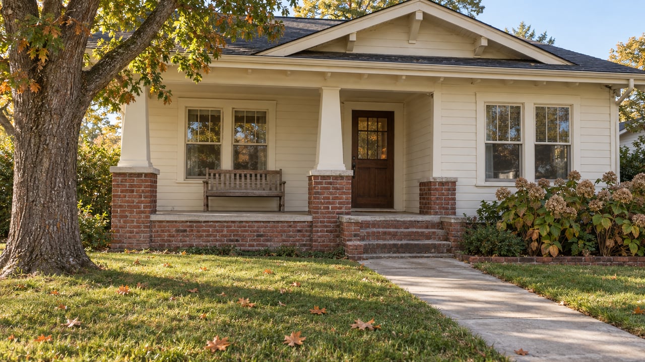 Cream-sided Craftsman home with a raised brick porch, mature oak and a concrete walk through an autumn lawn.
