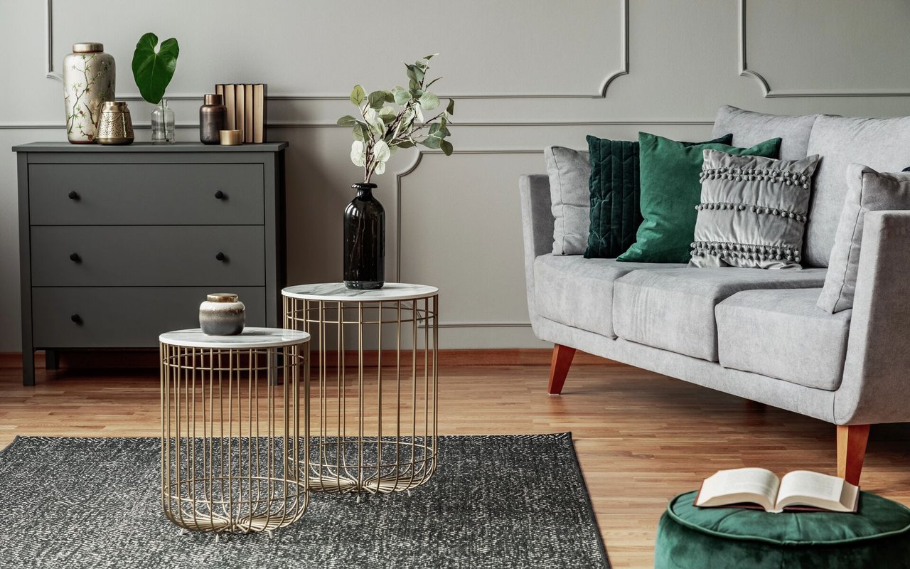 Living room with a gray sofa, emerald cushions, brass wire tables, and an open book on a green ottoman.