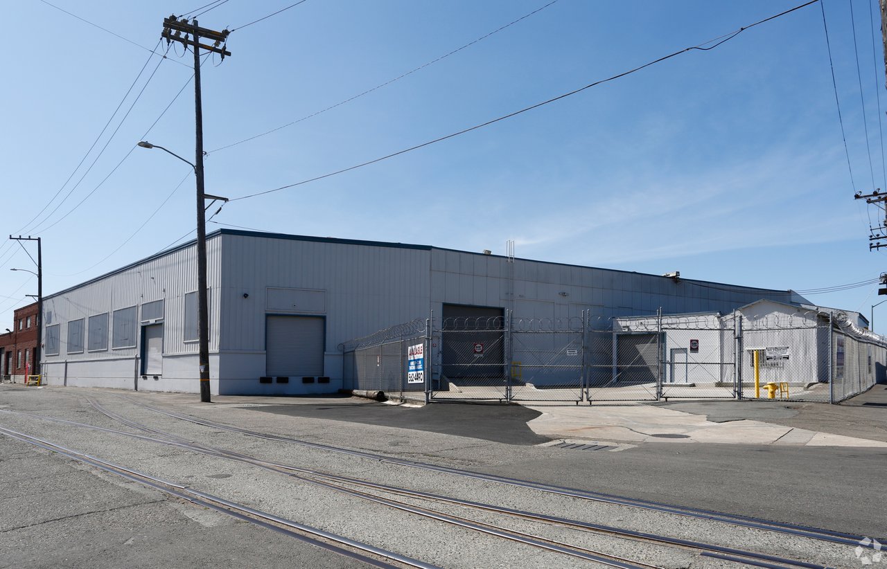 Industrial warehouse behind a razor-wire security fence, with railroad tracks crossing the foreground.