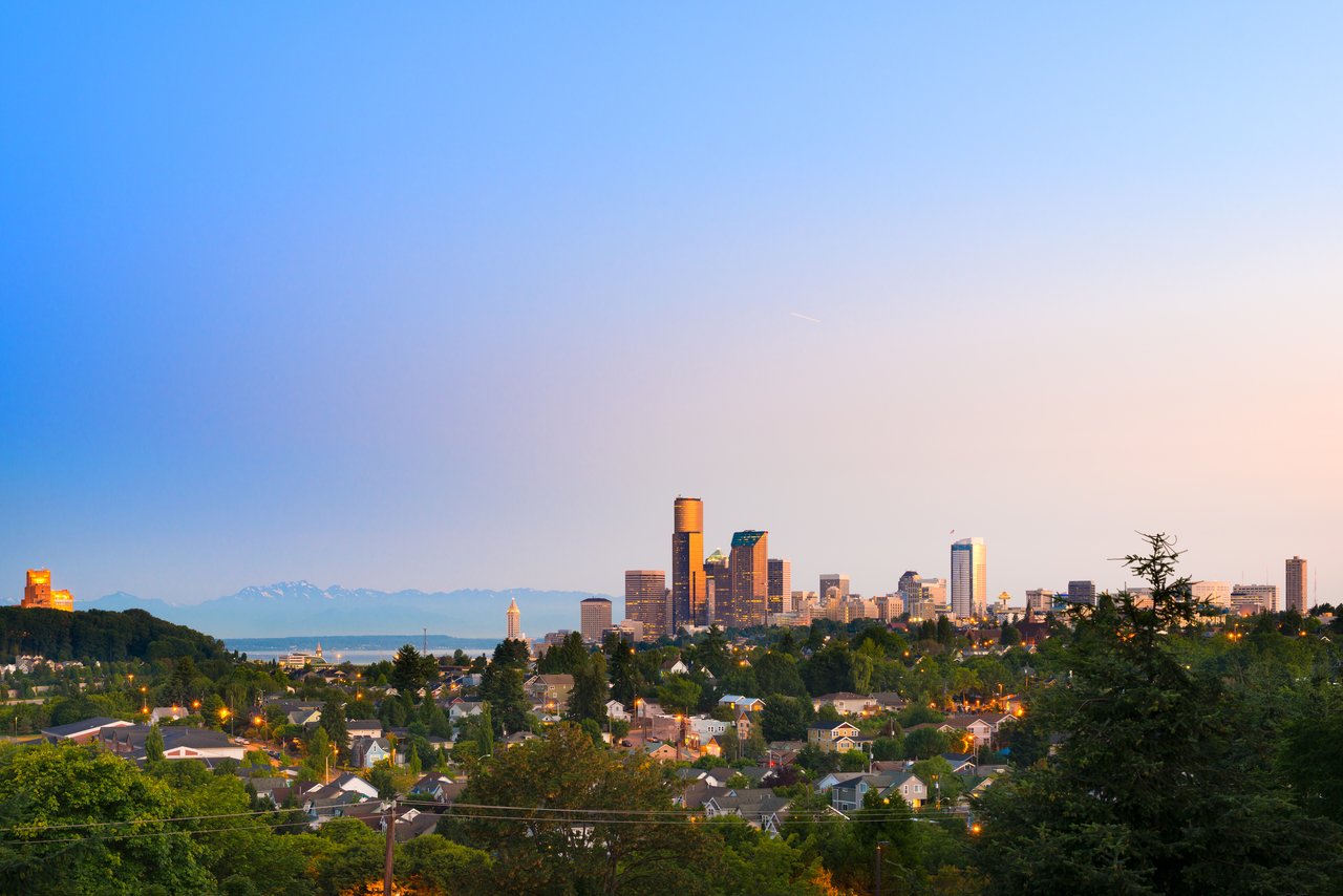 Seattle skyline at dusk above tree-lined neighborhoods, with mountains on the horizon.