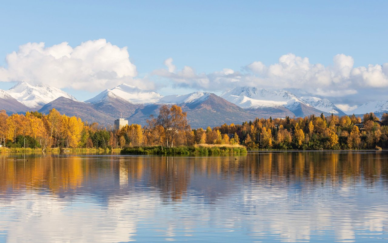 Golden autumn trees and snow-covered mountains reflected in an Anchorage lake.