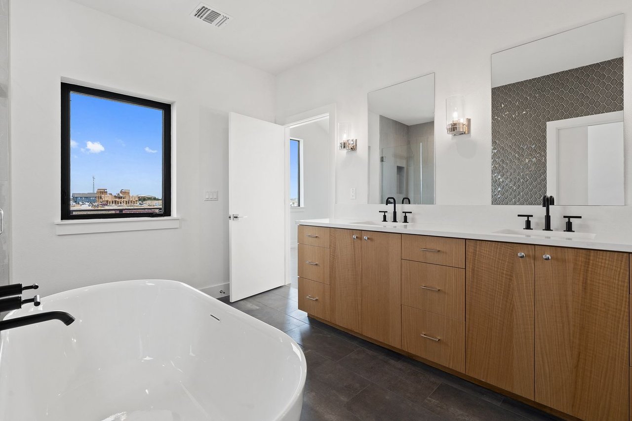 Modern bathroom with a freestanding soaking tub, double wood vanity, black faucets, and a window overlooking buildings.