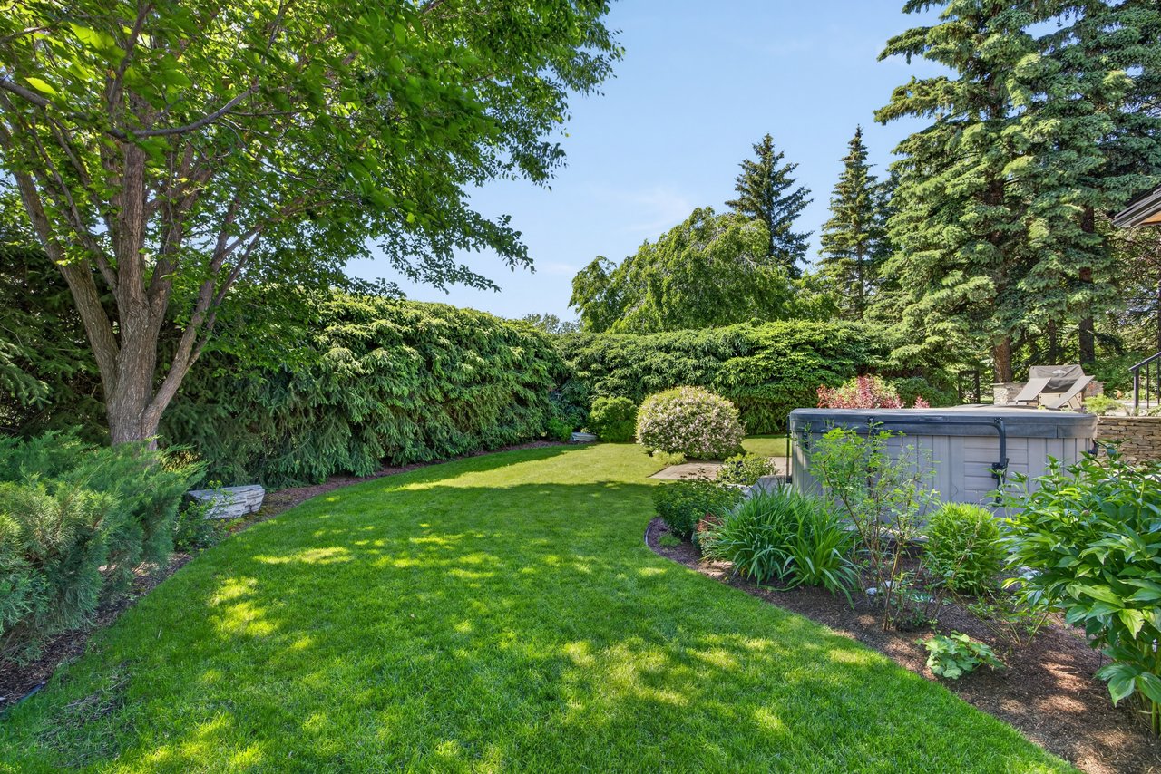 Landscaped backyard with a green lawn, mature trees, tall hedges, and an outdoor hot tub.