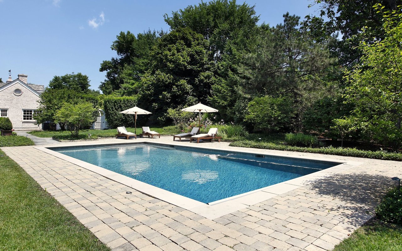 Backyard swimming pool with stone patio, lounge chairs, umbrellas, and mature trees.