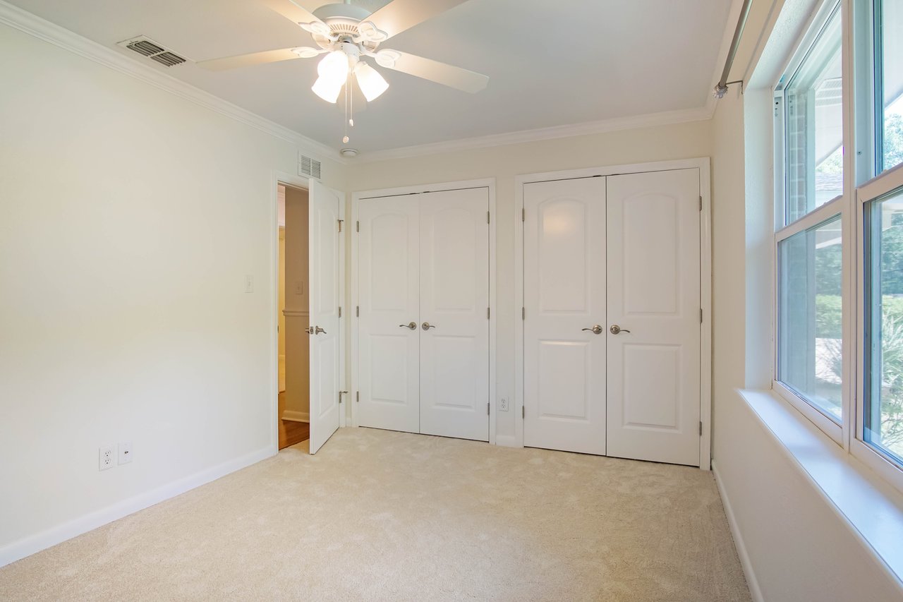 Spacious empty room with beige carpet, white walls, and ceiling fan. Features two closed double doors and large windows overlooking greenery.