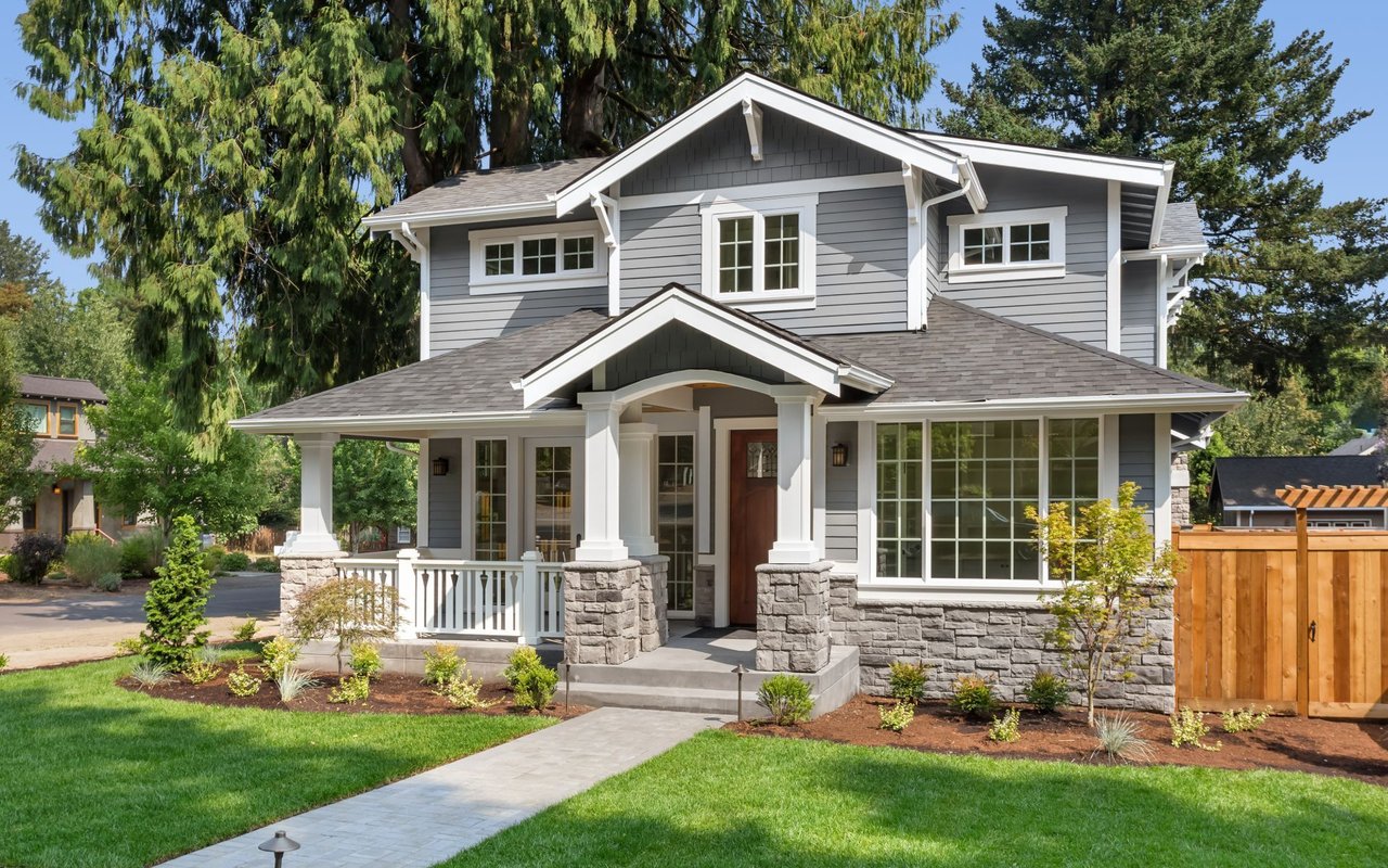 Two-story gray house with a covered porch, stone pillars, and a landscaped front lawn.