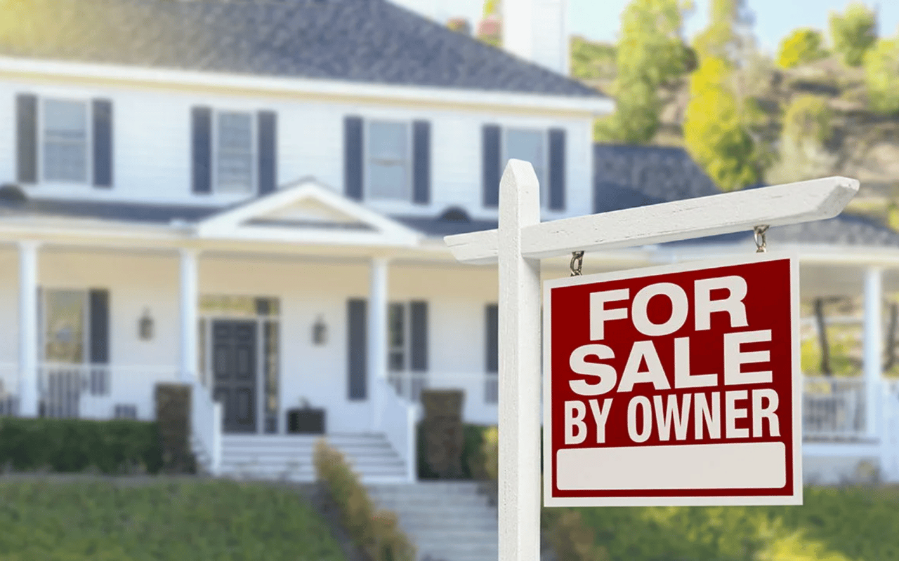 For-sale-by-owner sign in front of a white two-story house with dark shutters and a front porch.
