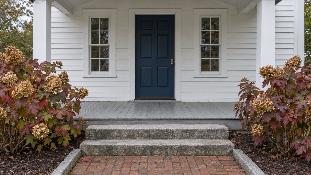 White clapboard home entry with a navy door, paired windows, weathered porch boards, granite steps and burgundy oakleaf hydrangeas.