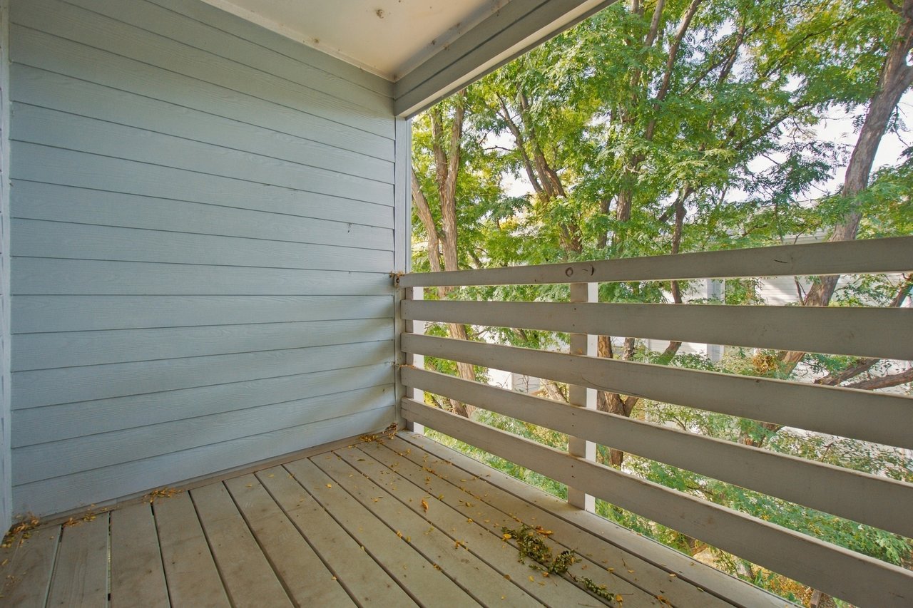 Empty apartment balcony with gray plank flooring and leafy trees beyond the railing.