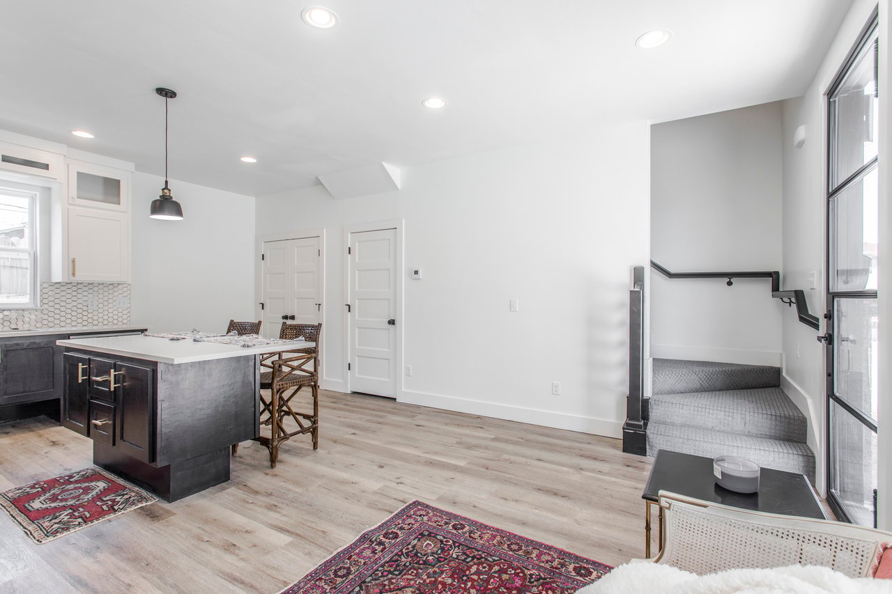 Open kitchen and living area with a dark island, patterned rugs, and carpeted stairs beside a glass door.